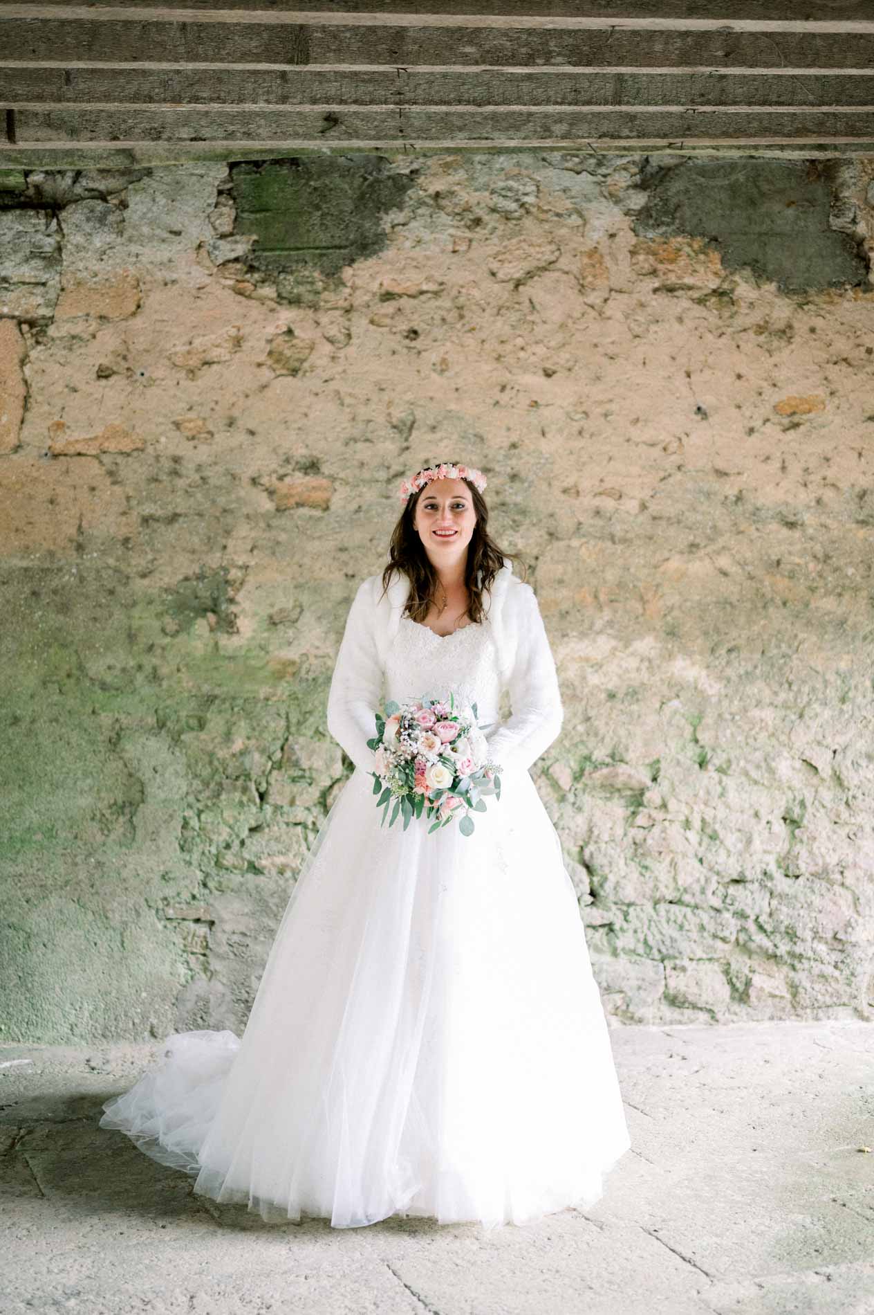 Bride in a ball gown and white wrap standing before an old wall