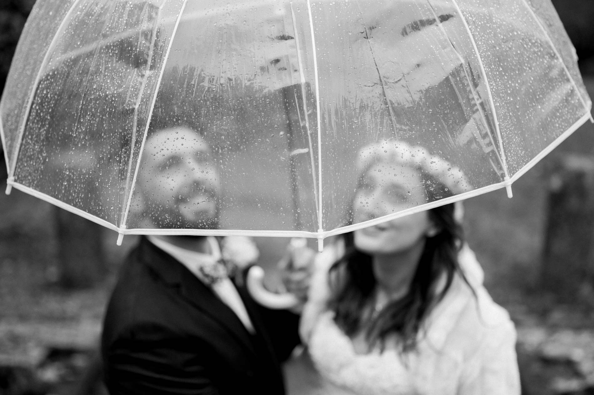 Bride and groom under a clear umbrella looking up at the rain, in black and white
