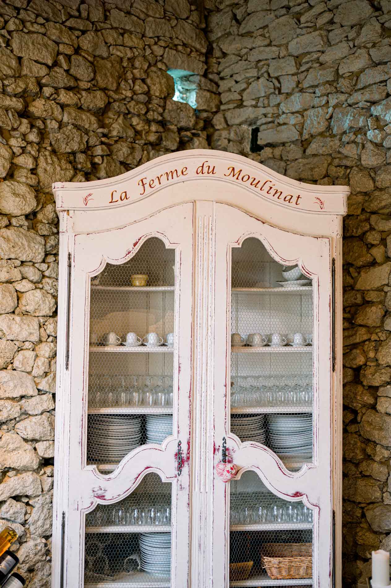 Antique white dresser marked La Ferme du Moulinat against a stone wall