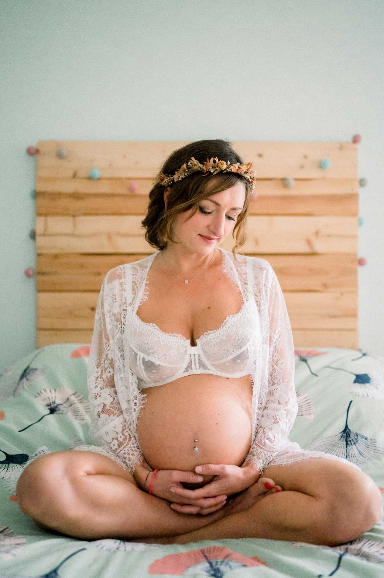 Future maman assise en tailleur sur le lit, mains en cœur sur le ventre