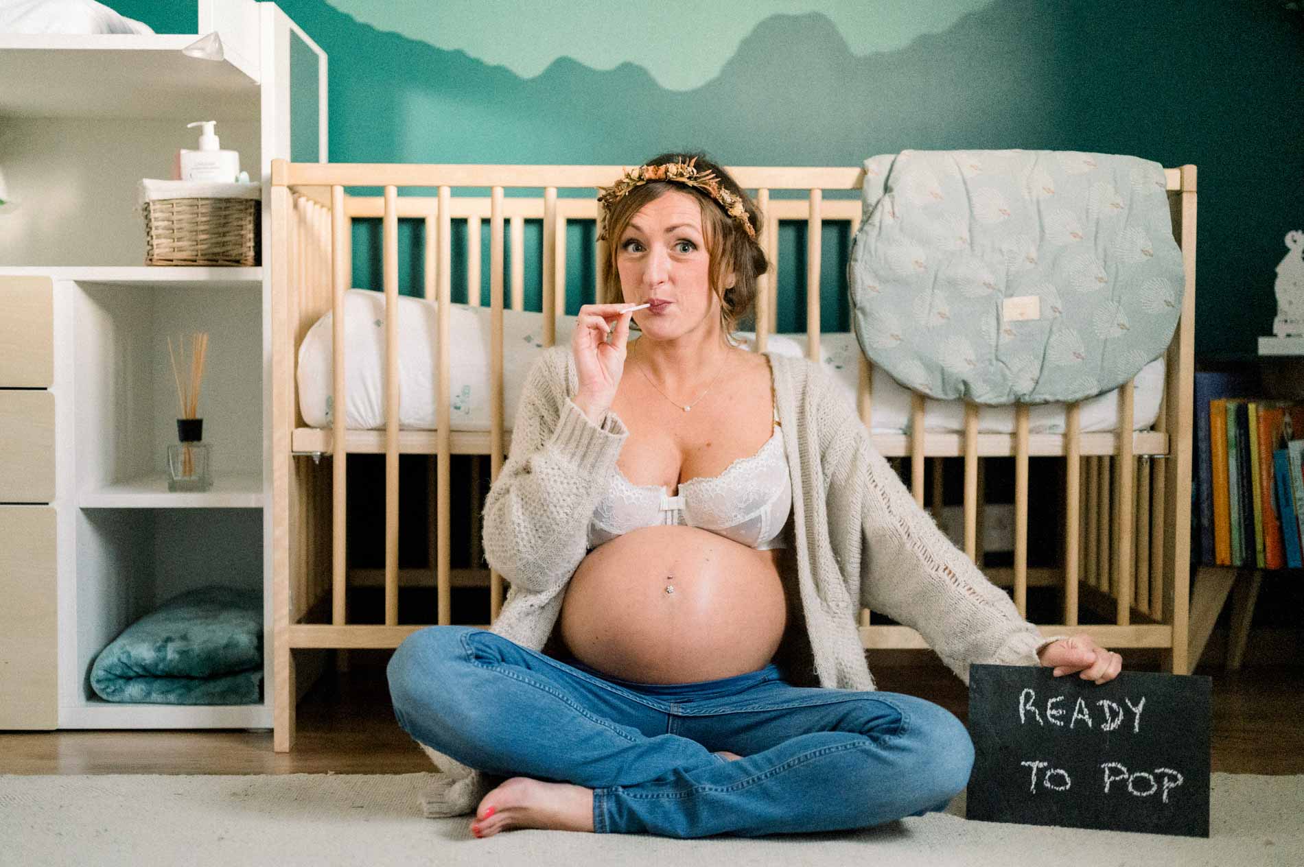 Future maman devant un berceau avec une ardoise « Ready to pop », séance grossesse à domicile