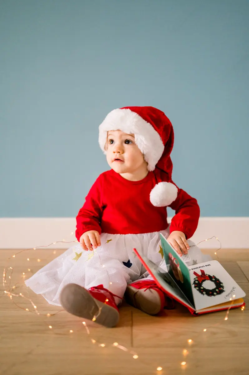 Baby in a Santa hat sitting on the floor with a book and fairy lights
