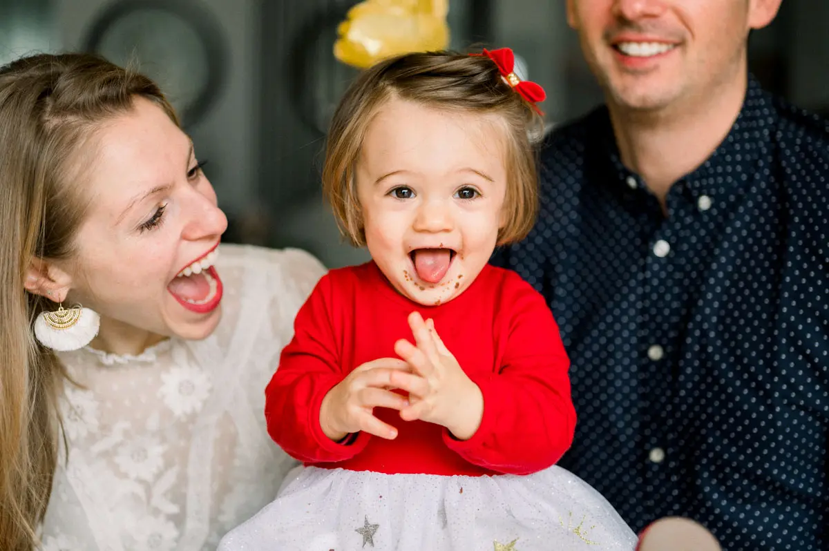 Little girl in a red jumper laughing between her parents on her first birthday