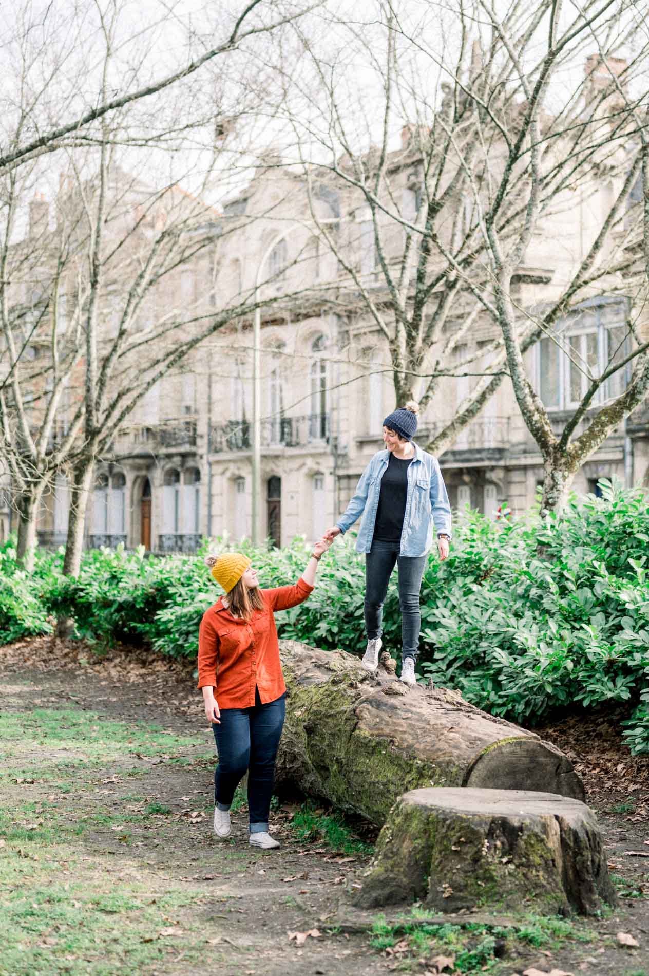 She helps her partner down from a log, Bordeaux facades behind