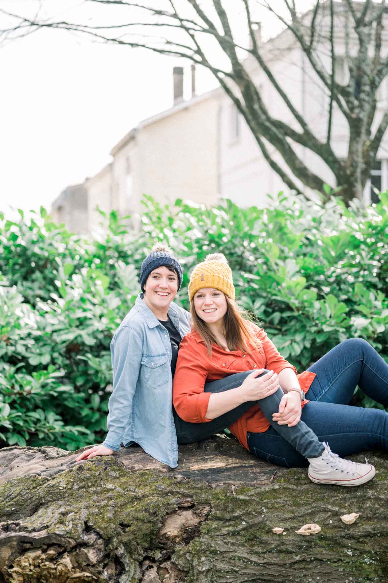 Two women sitting on a log, smiling, in front of lush greenery