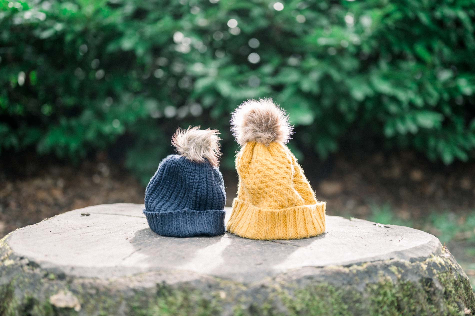Two pompom beanies, mustard and navy, resting on a tree stump