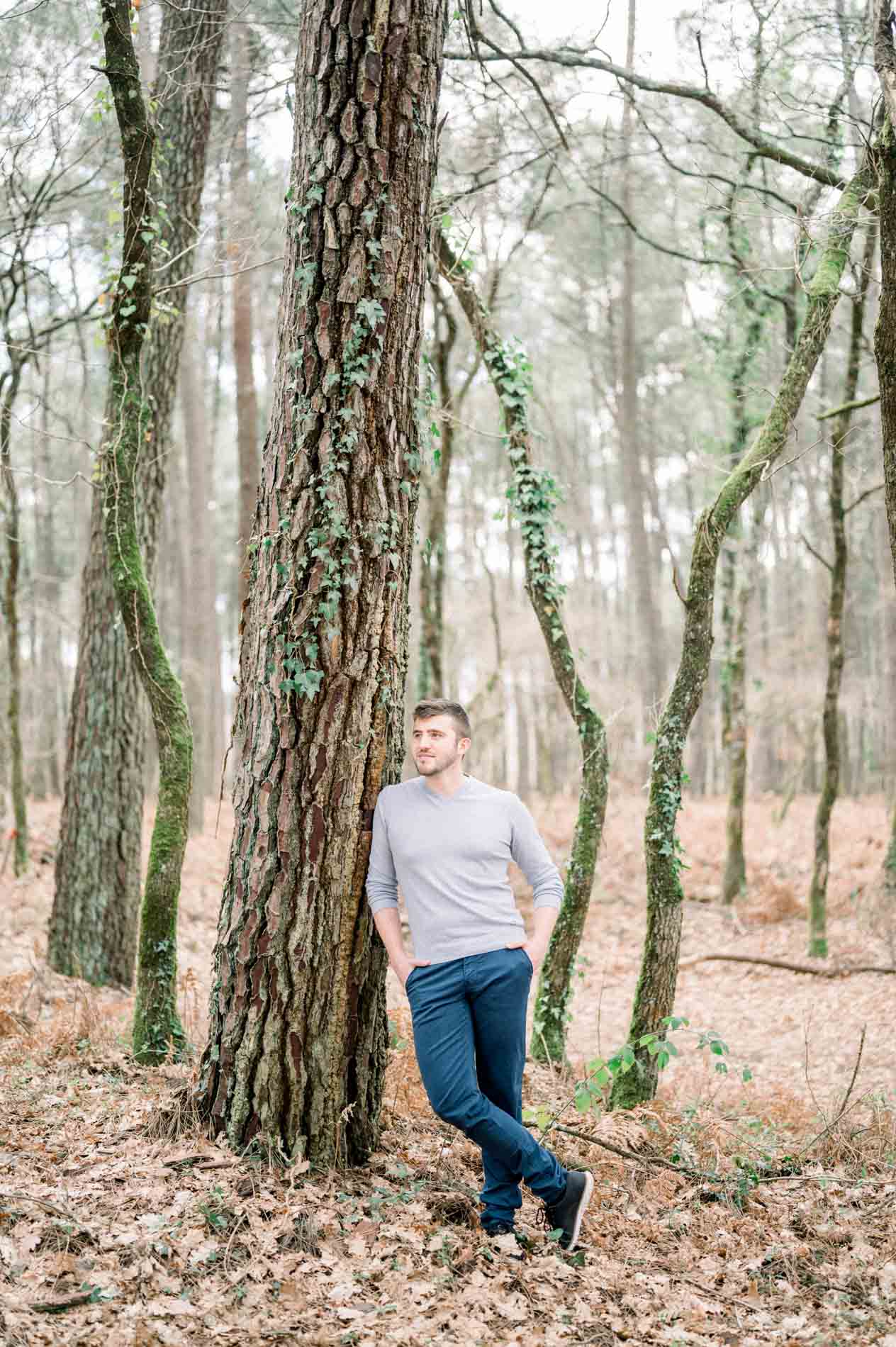 Man in a grey jumper leaning against a pine trunk in the forest