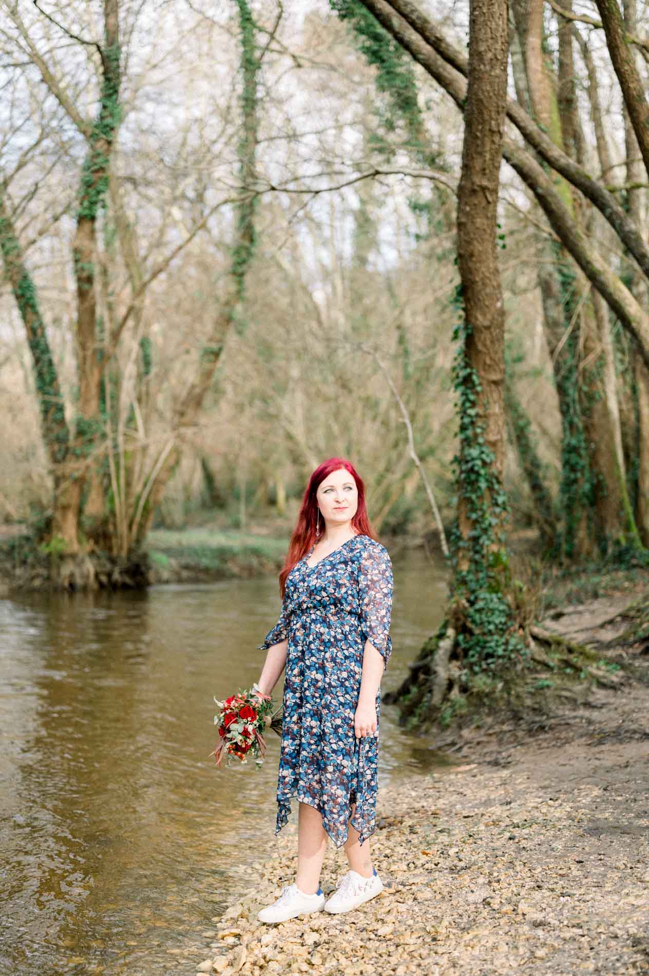 Woman in a floral dress by a forest stream, bouquet in hand