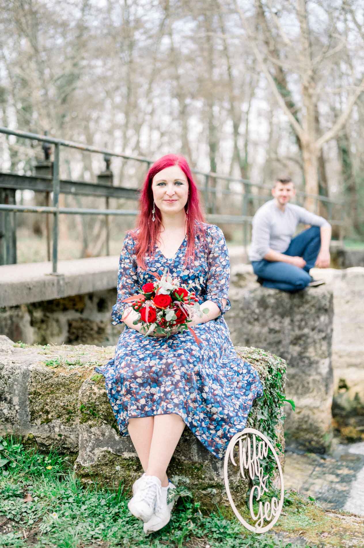 Woman sitting with her bouquet by a weir, her partner crouching behind