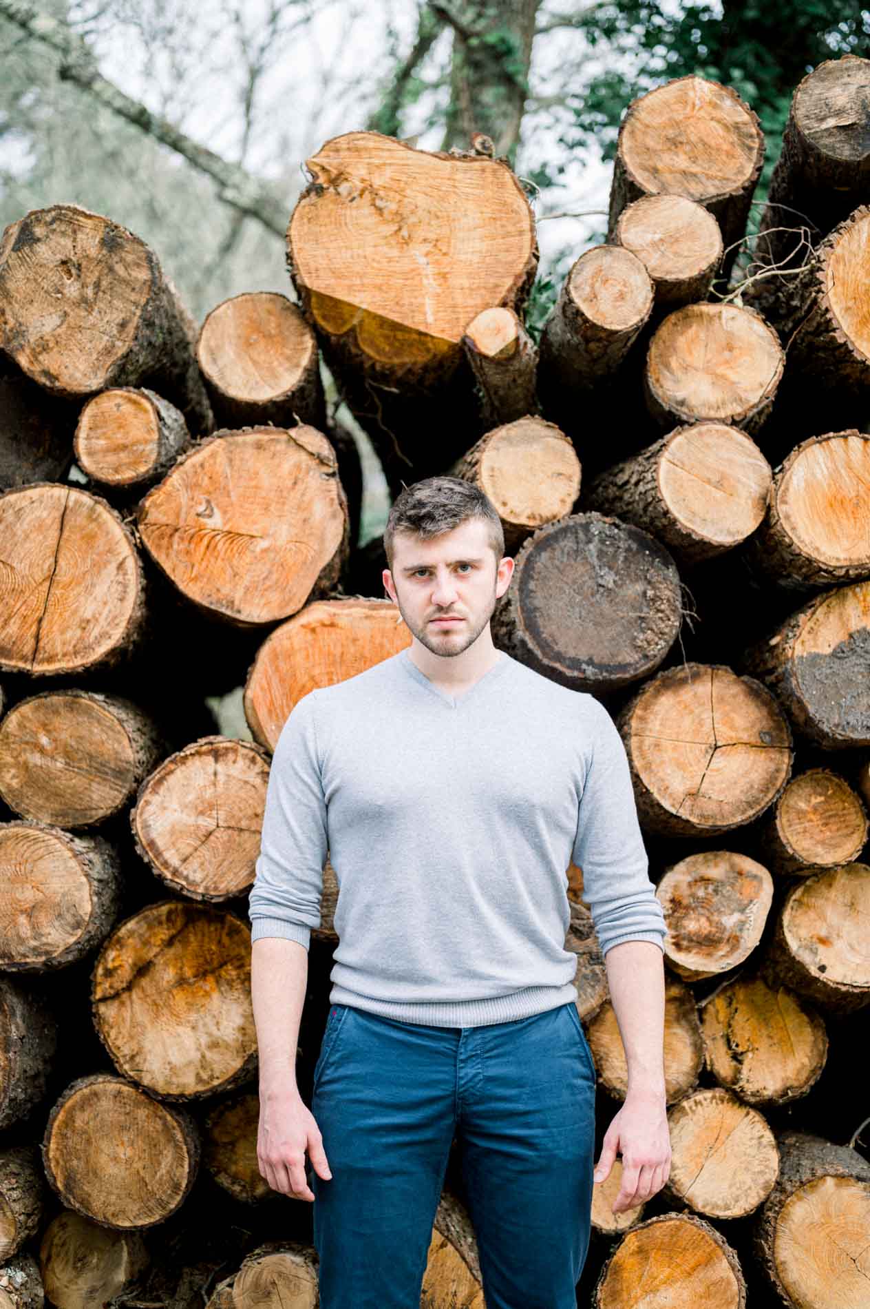 Man in a grey jumper in front of a stack of cut logs