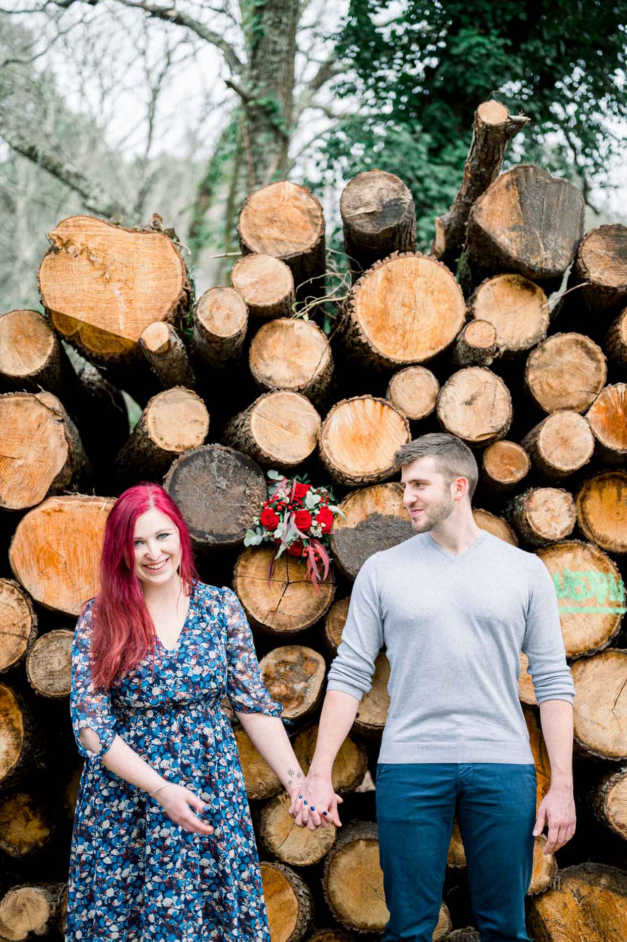 Smiling couple holding hands by a log pile, bouquet of roses