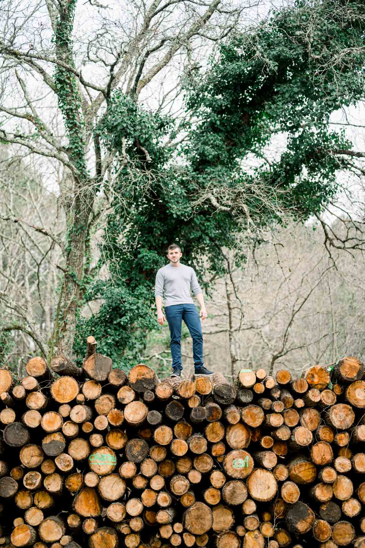 Man standing on top of a large log pile beneath the trees