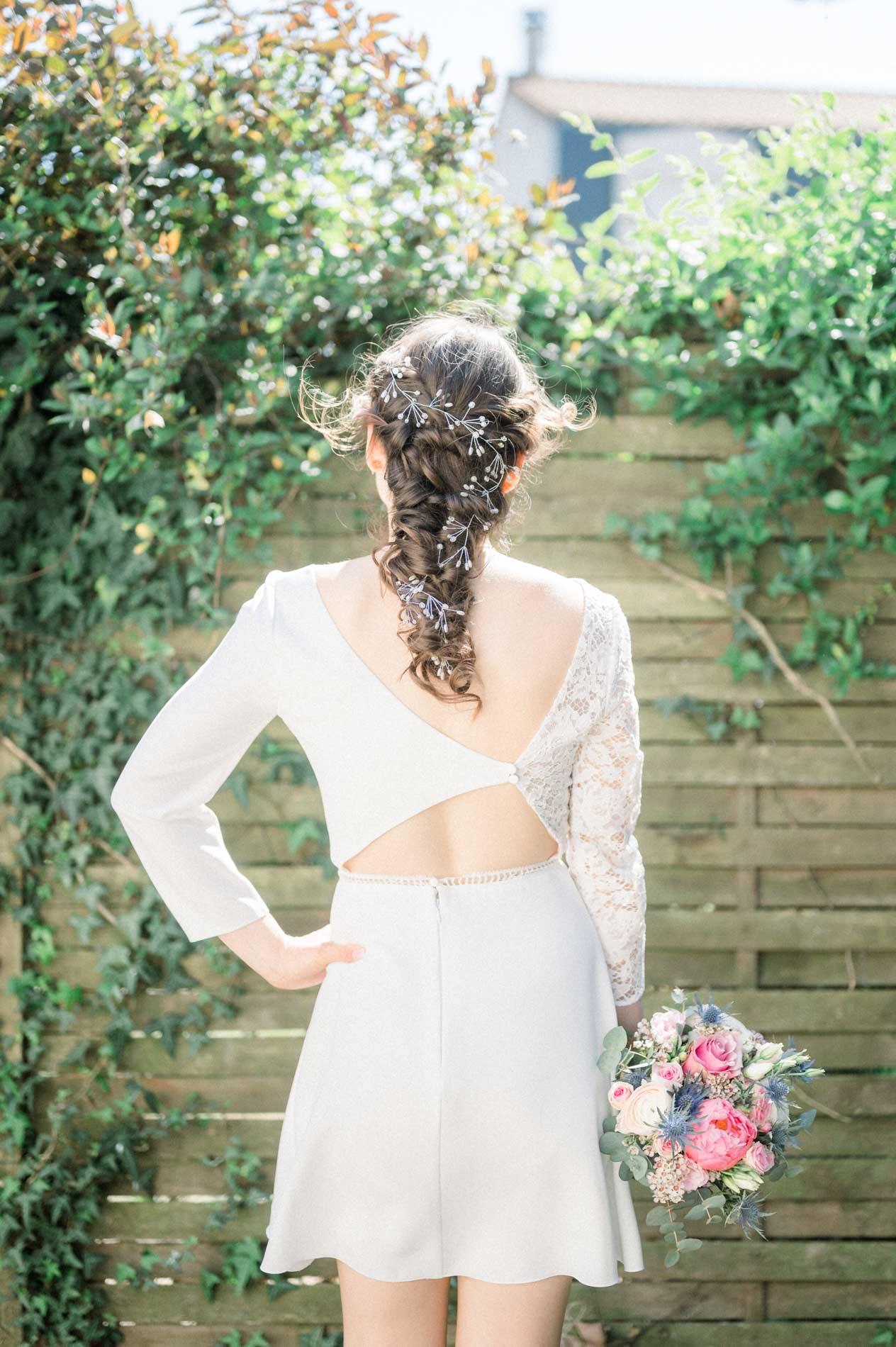 La mariée de dos, robe à dos ouvert et bouquet, devant une palissade fleurie