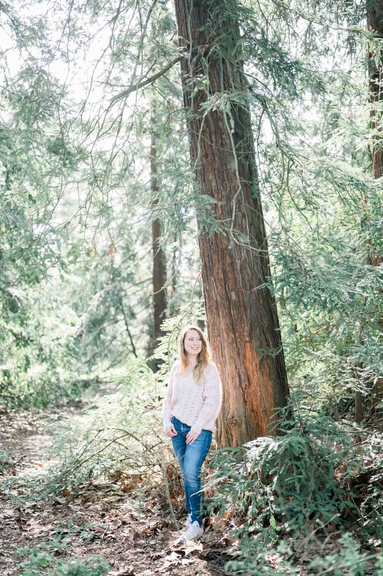 The sister in a pale pink jumper standing at the foot of a redwood in the woods