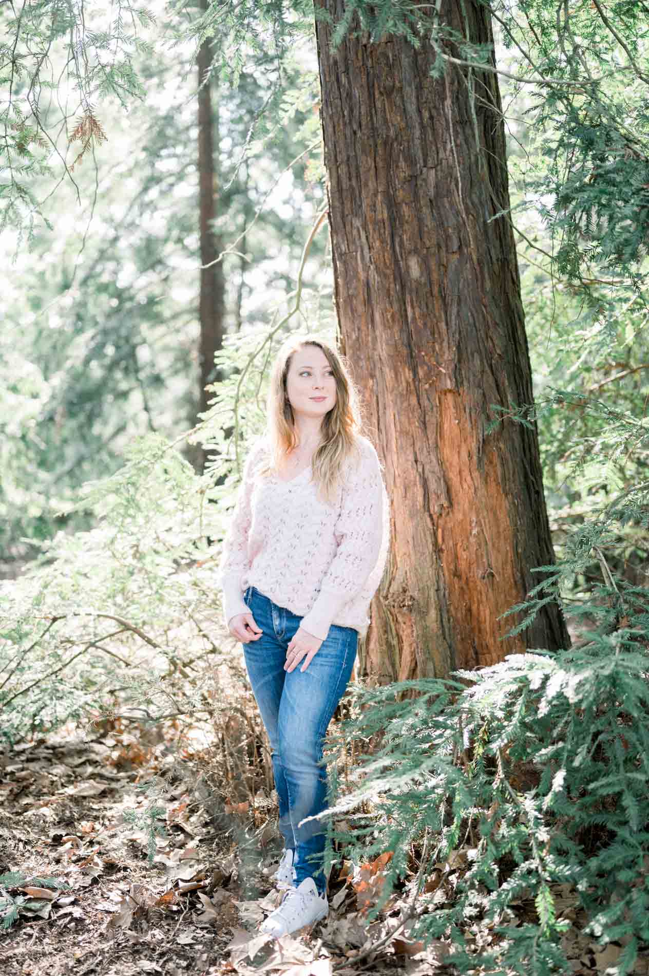 The sister in jeans and a light jumper leaning against a large tree trunk
