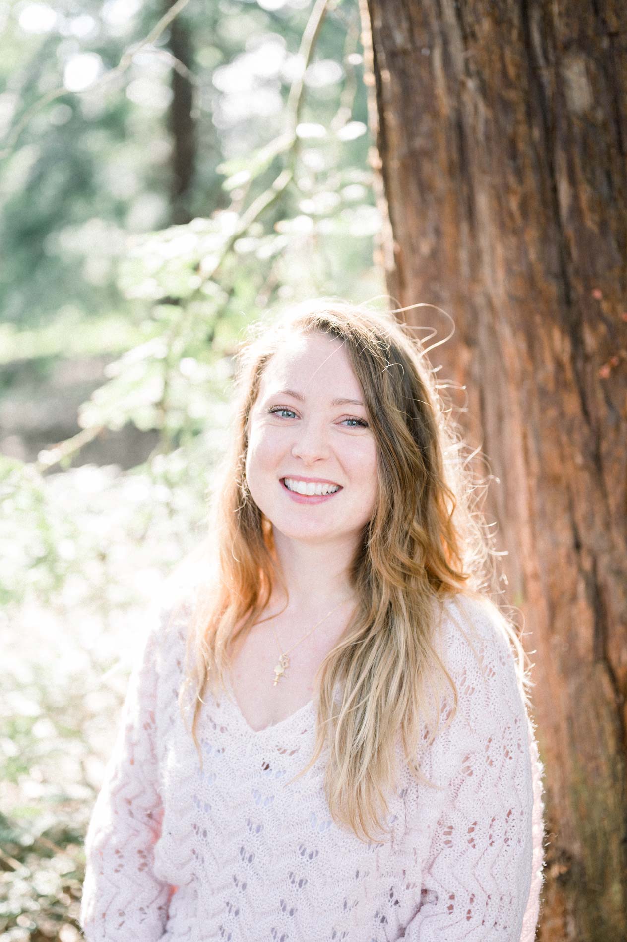 Smiling portrait of the sister with long blonde hair in front of a reddish trunk