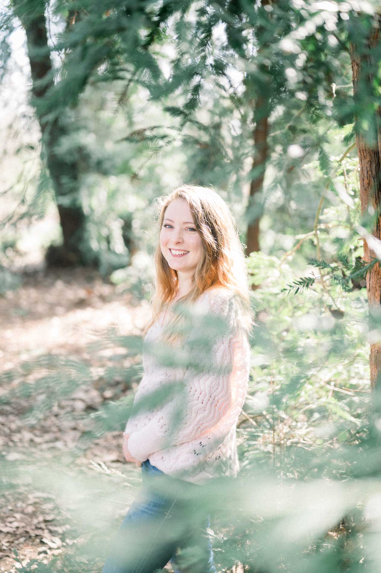 The sister smiling through conifer branches in the foreground