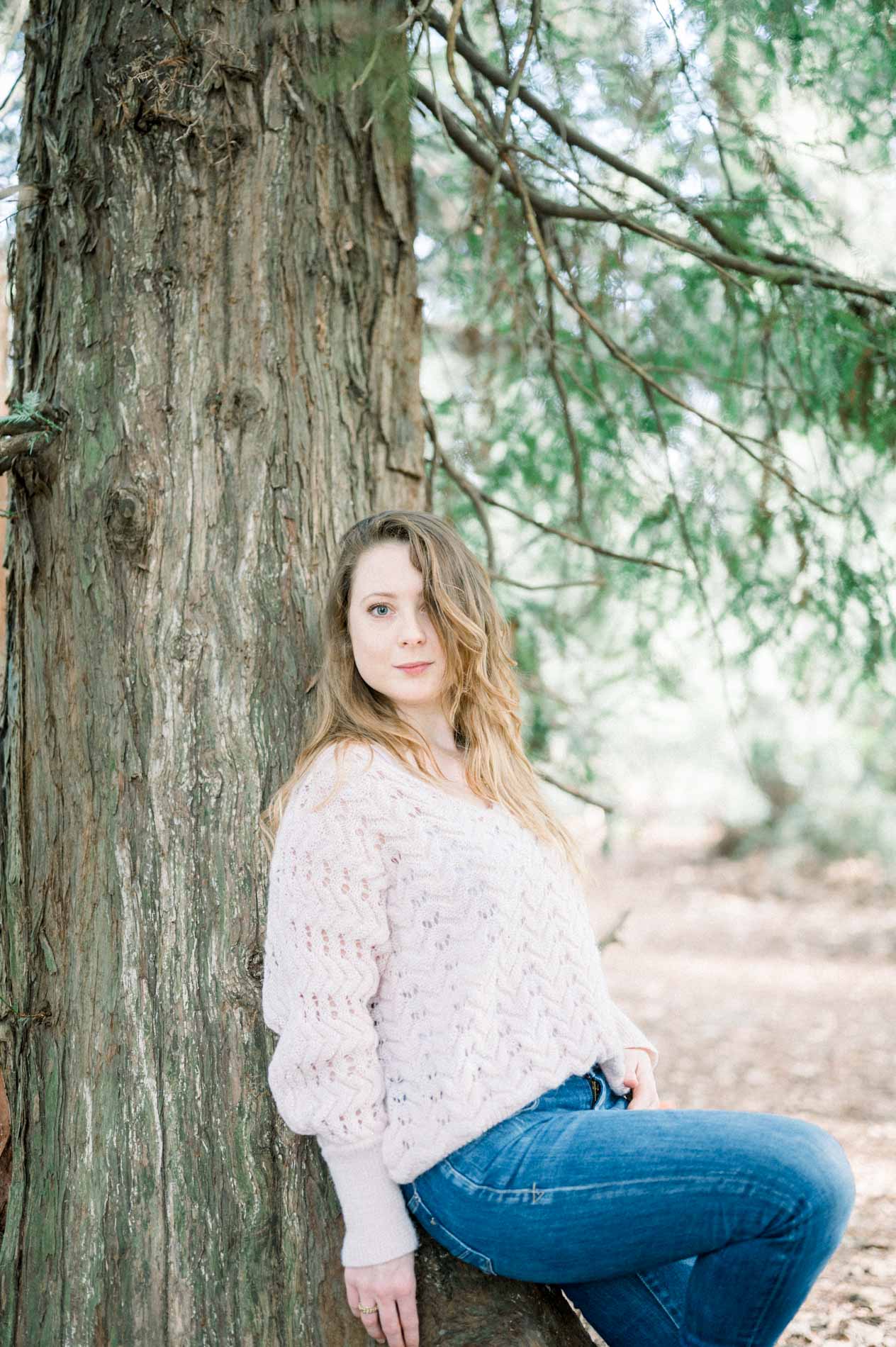 The sister in a wool jumper sitting against a tree, looking at the camera