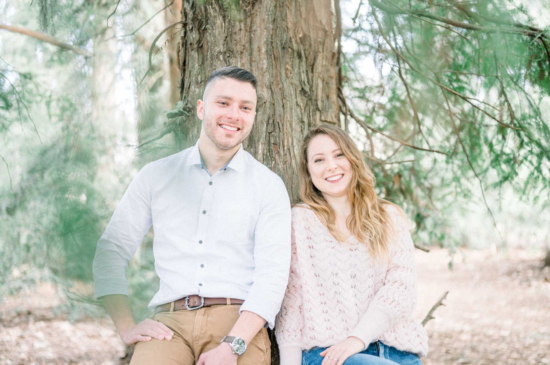 Brother and sister sitting side by side at the foot of a tall tree, smiling
