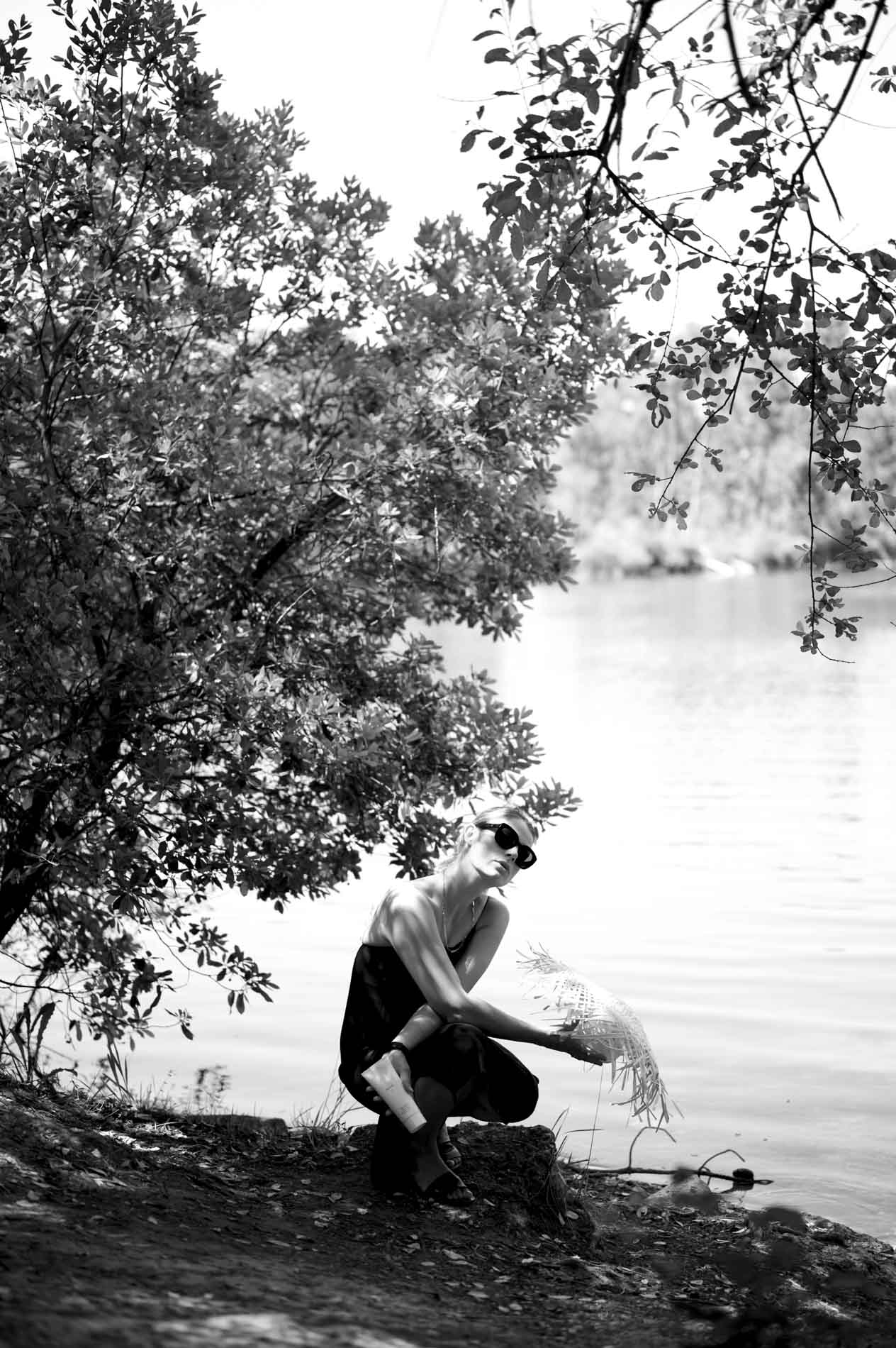 Model crouching under a tree at a lakeside, black and white