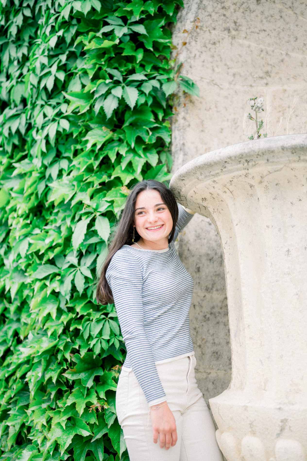 Jeune femme souriante adossée à un vase en pierre devant un mur de vigne vierge