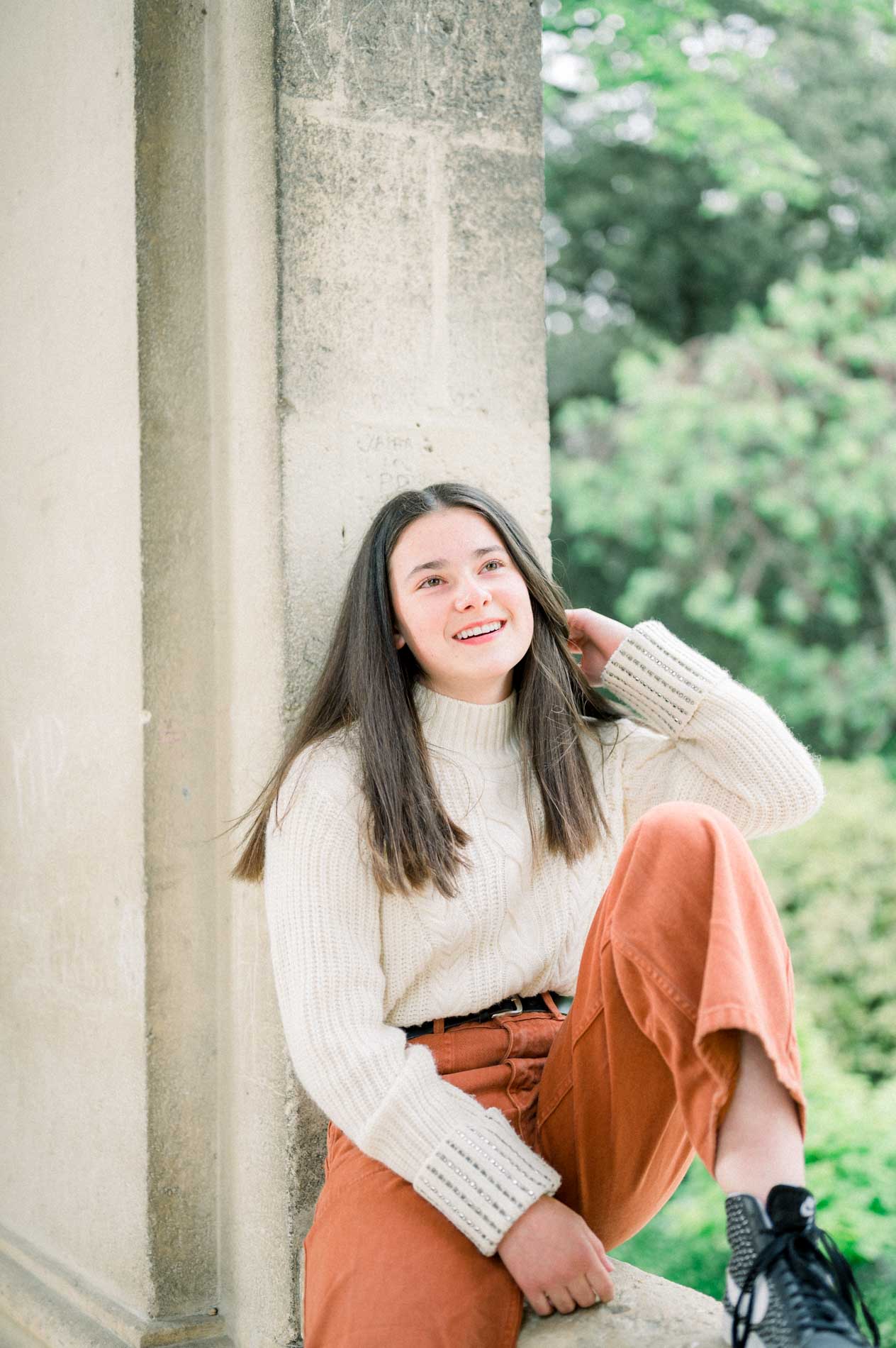 Jeune femme en pull crème assise contre une colonne en pierre, sourire aux lèvres