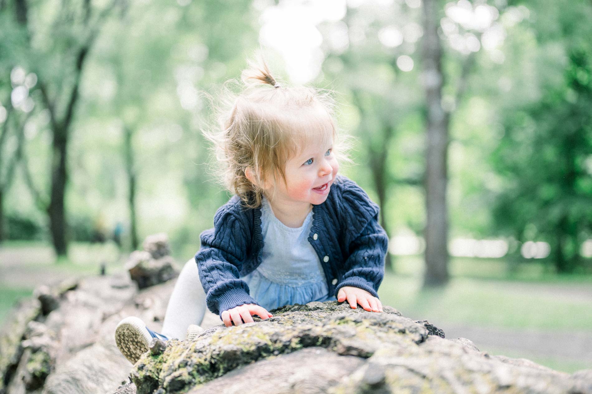 The little blonde girl smiling as she climbs onto a tree trunk