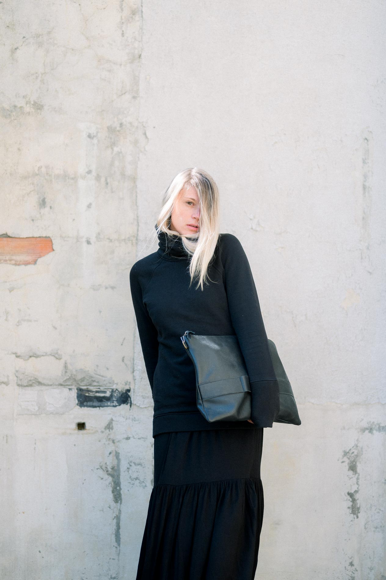 Model in a black outfit with a leather bag against a weathered wall