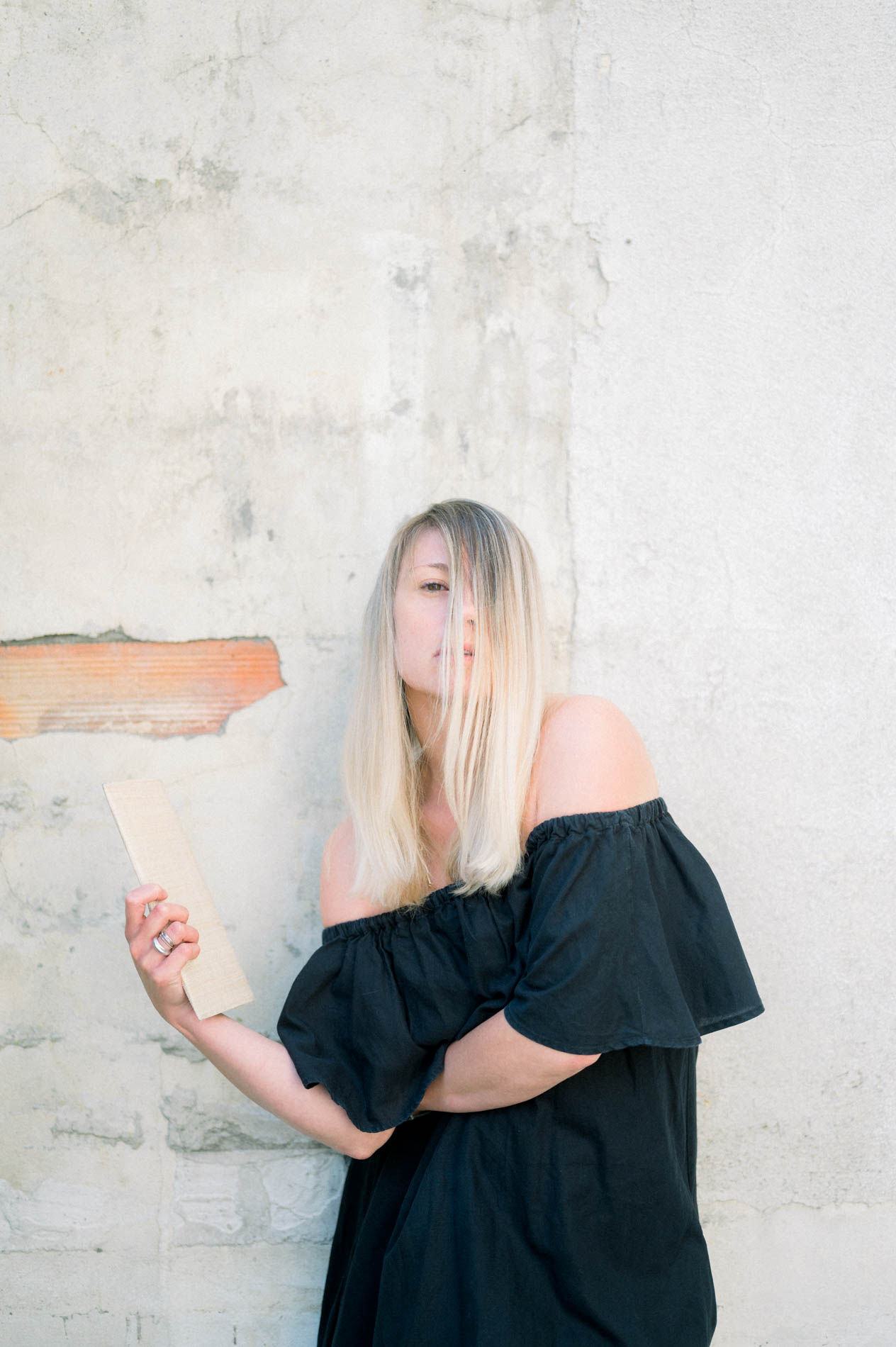 Model in an off-shoulder black dress holding a book against a pale wall
