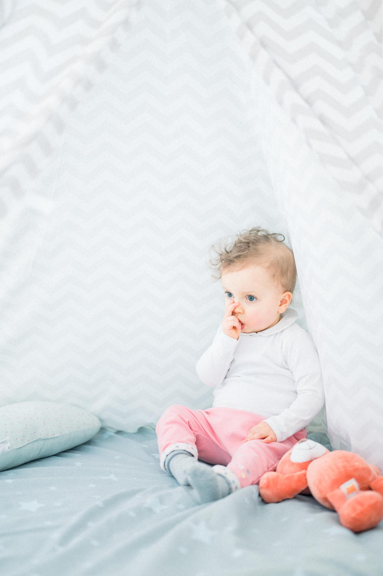 Baby sitting in a teepee, finger in mouth, beside a toy fox