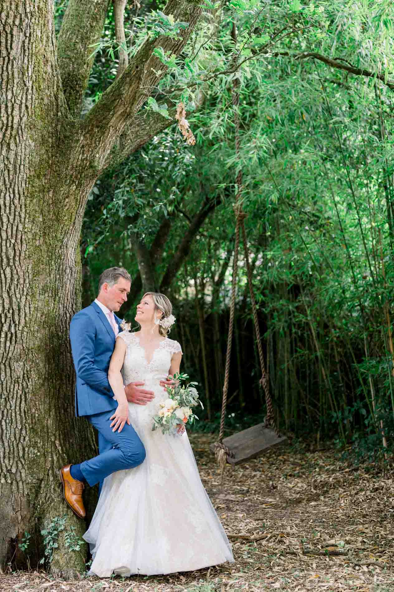 Groom leaning against a large tree, holding the bride in the grounds