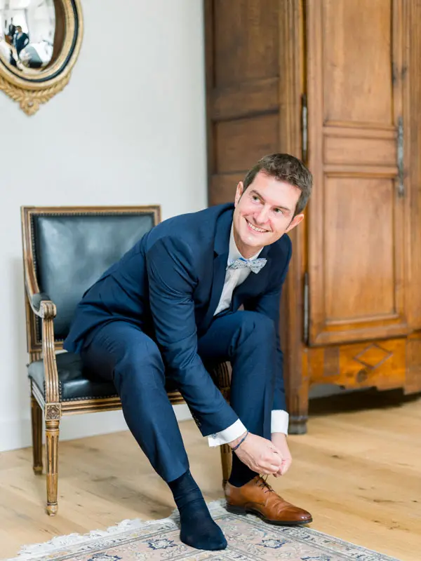 Groom in a blue suit tying his shoes, seated in an antique armchair