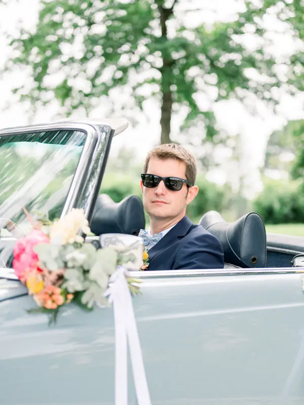 Groom in sunglasses at the wheel of the flower-decked vintage convertible