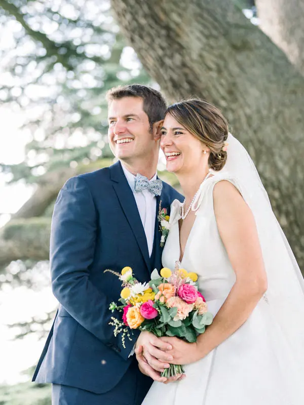 Smiling couple forehead to forehead under the cedar, bright bouquet in hand