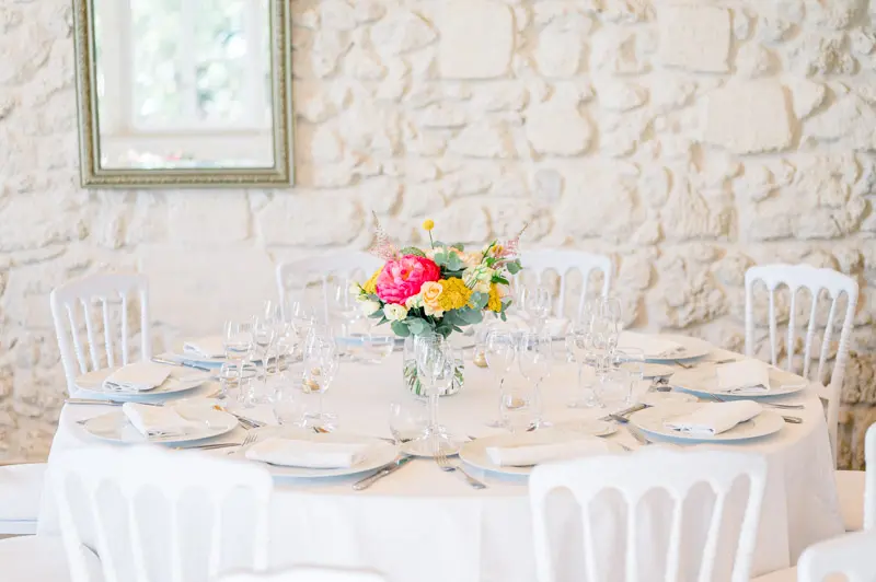 Round table with white linen and a colourful centrepiece against a stone wall