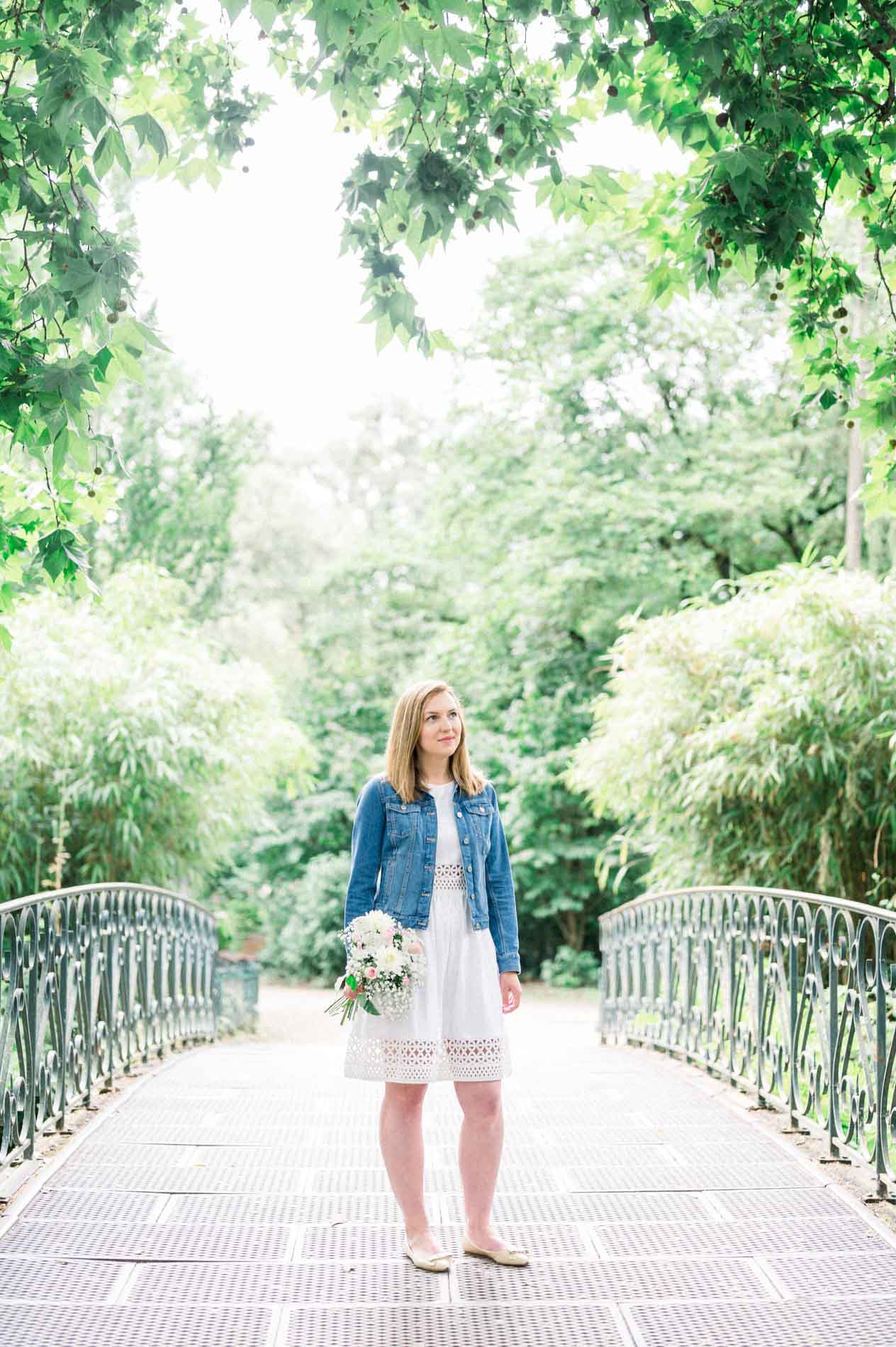 Tamara in a white dress and denim jacket on a footbridge in the Jardin Public, bouquet in hand
