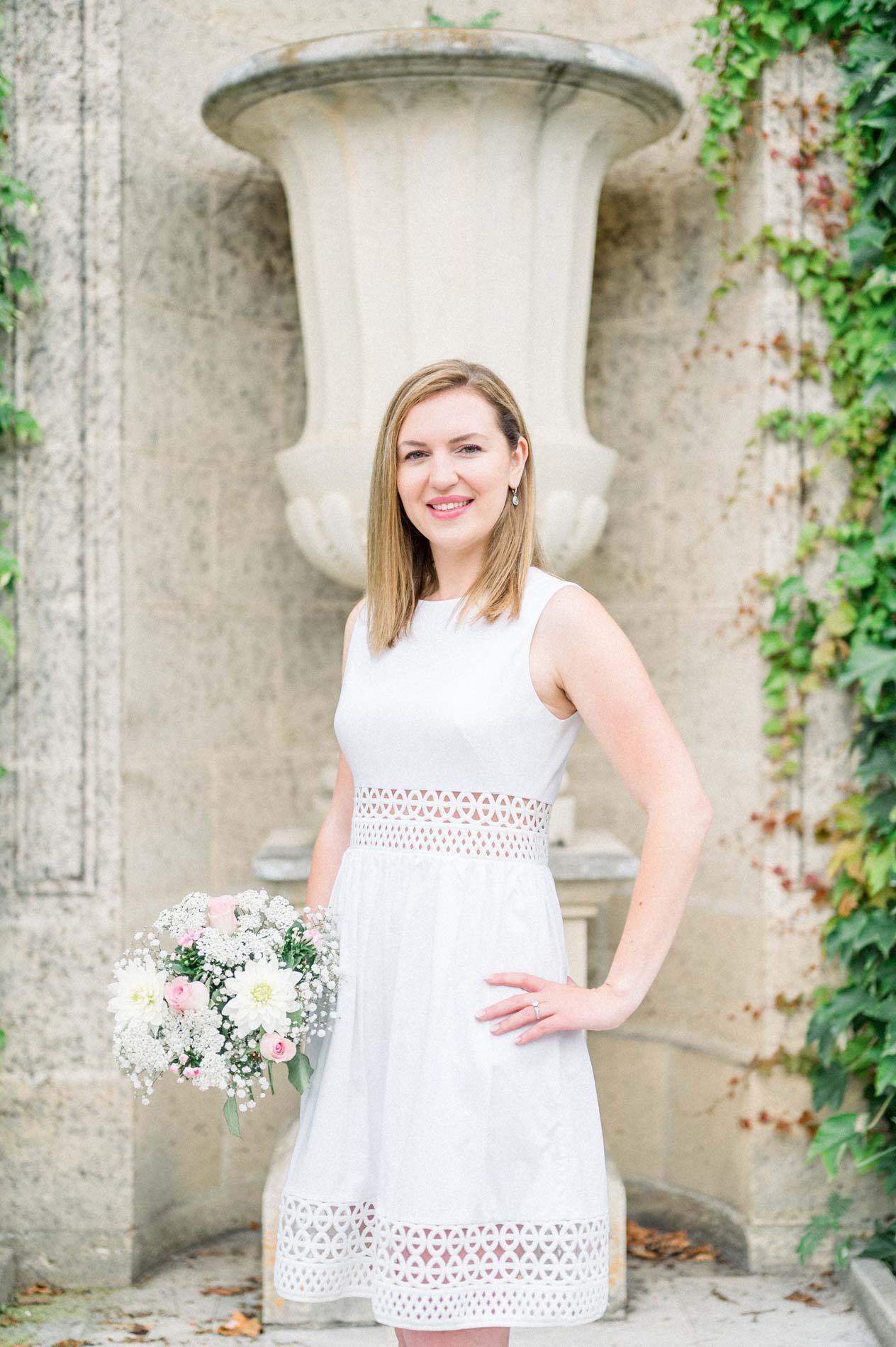 Tamara in a white dress holding her bouquet in front of a stone urn