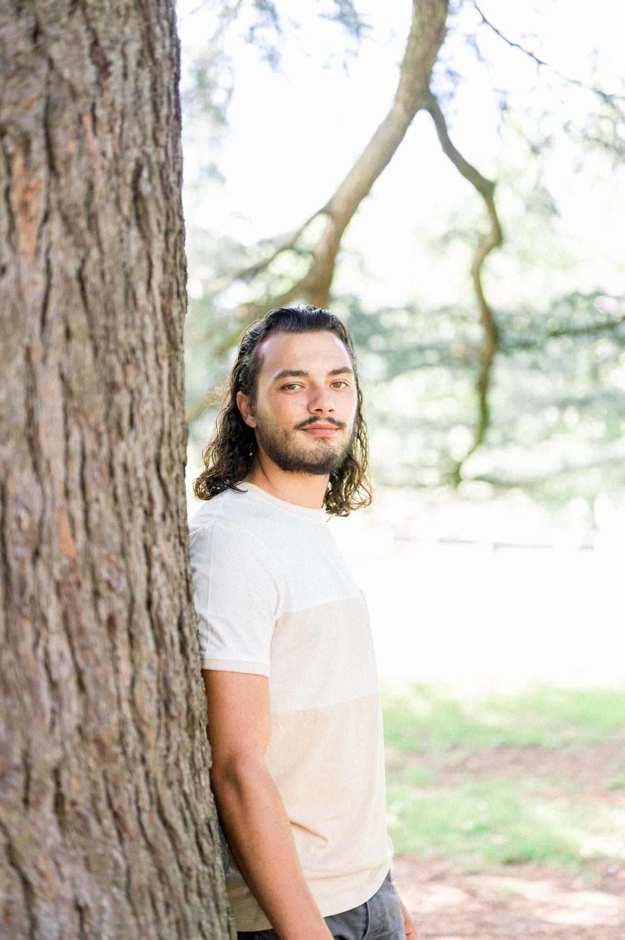 Young man with long hair leaning against the trunk of a tall tree