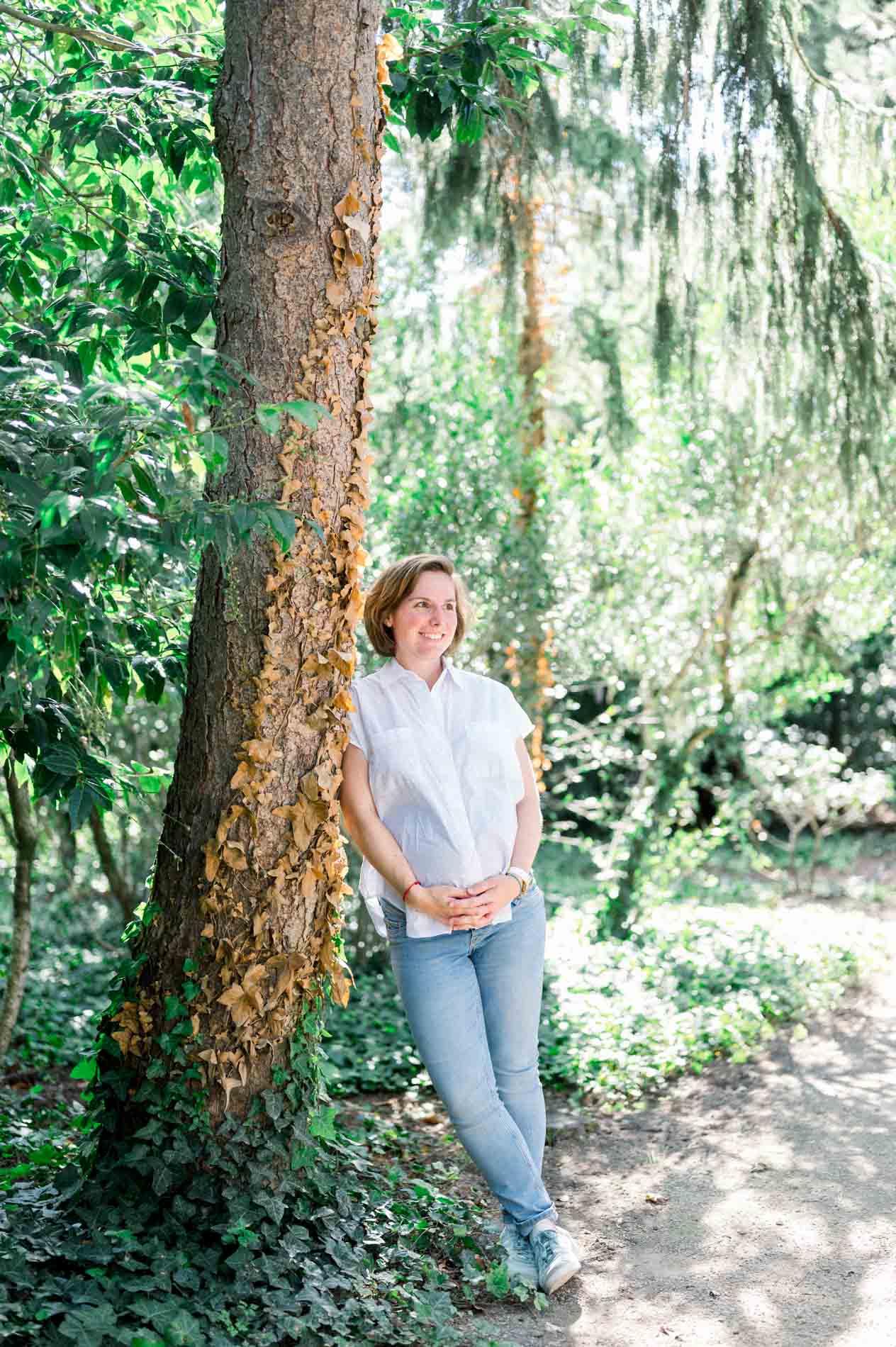 Pregnant woman leaning against a tall tree, hands on her belly