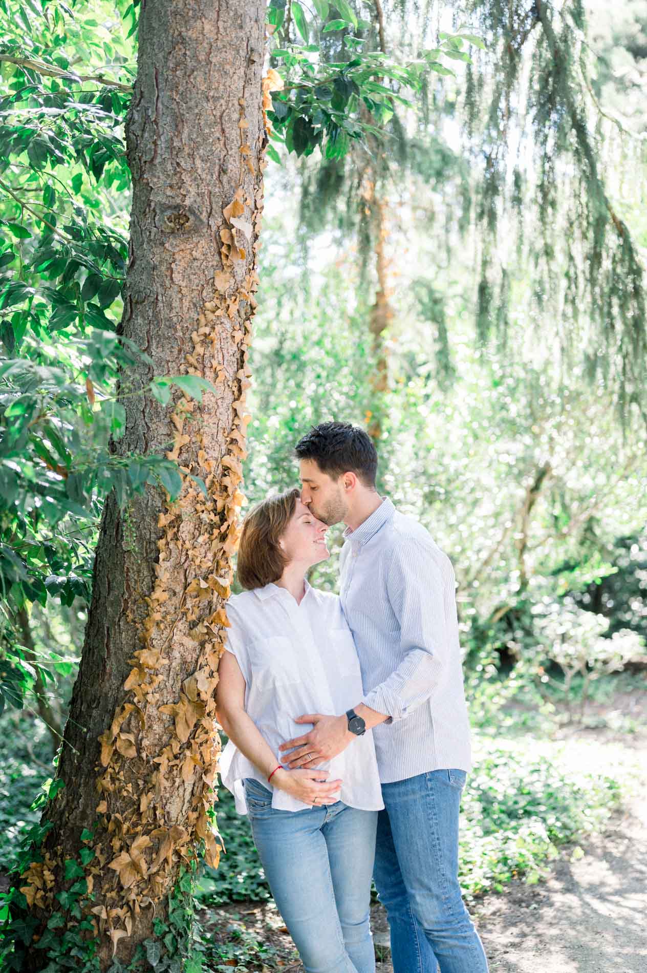 Couple expecting a baby nestled against a tree trunk in the park