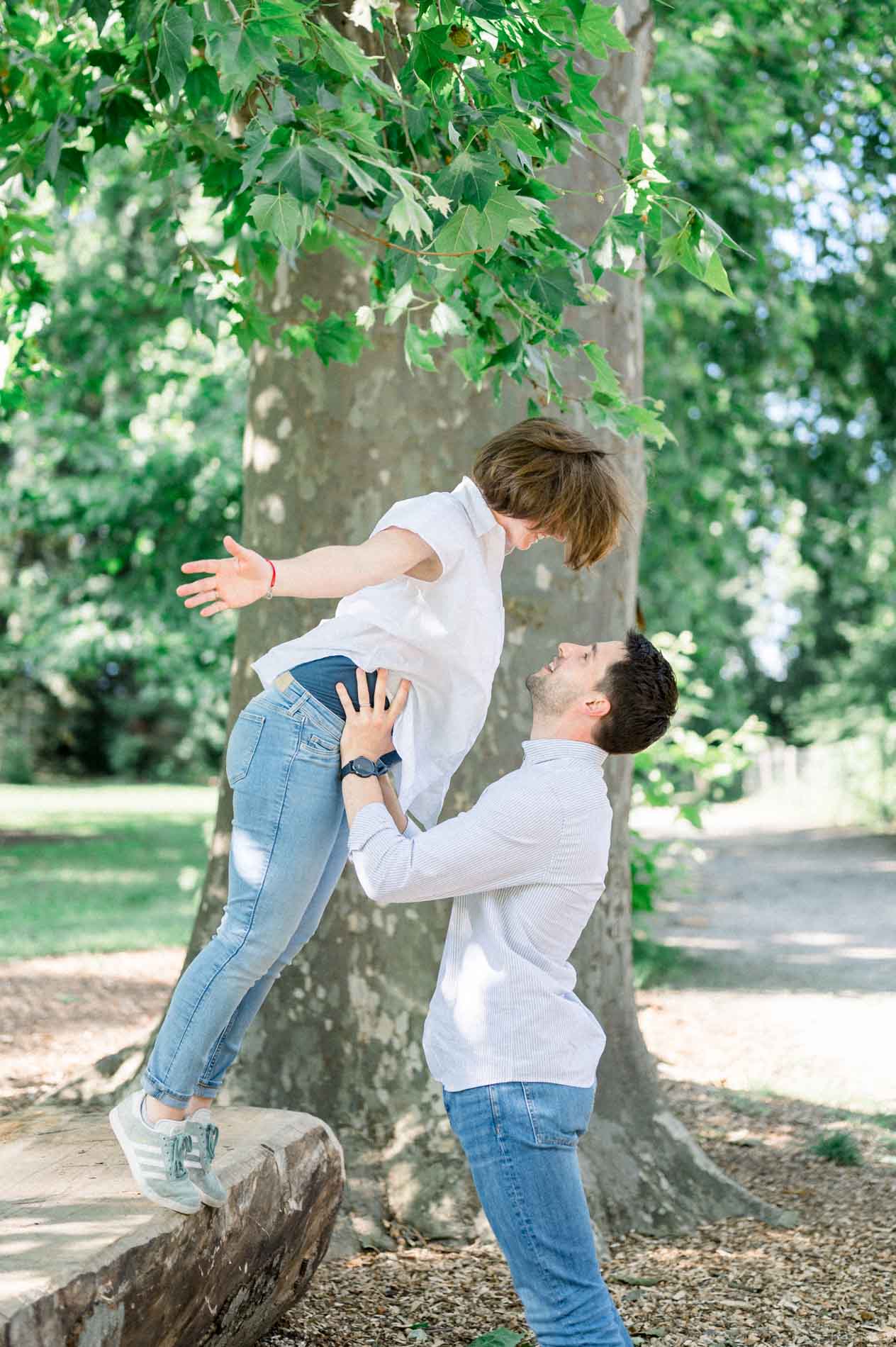 Father-to-be joyfully lifting his pregnant partner under a plane tree