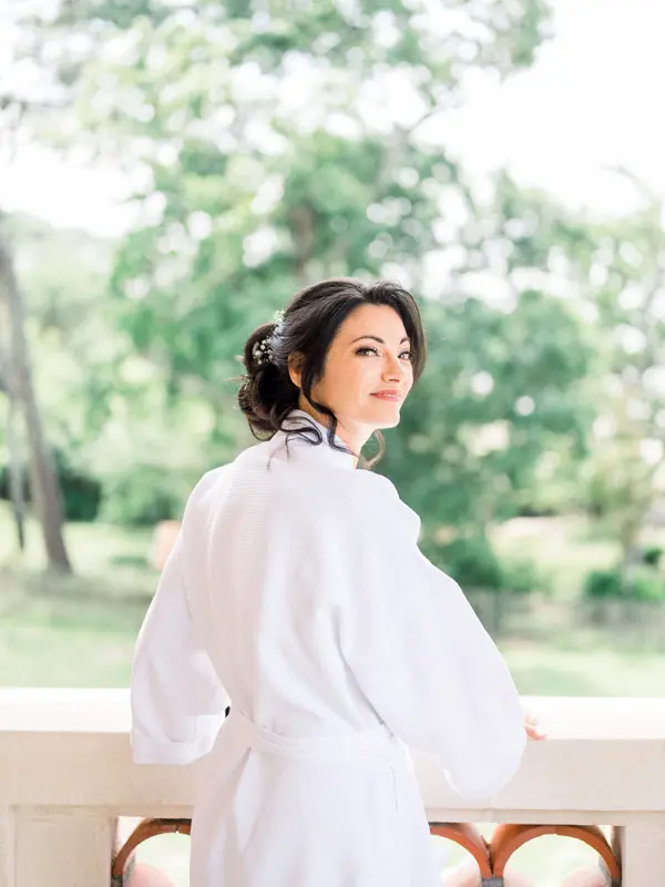 The bride in a white robe glancing back from the villa balcony