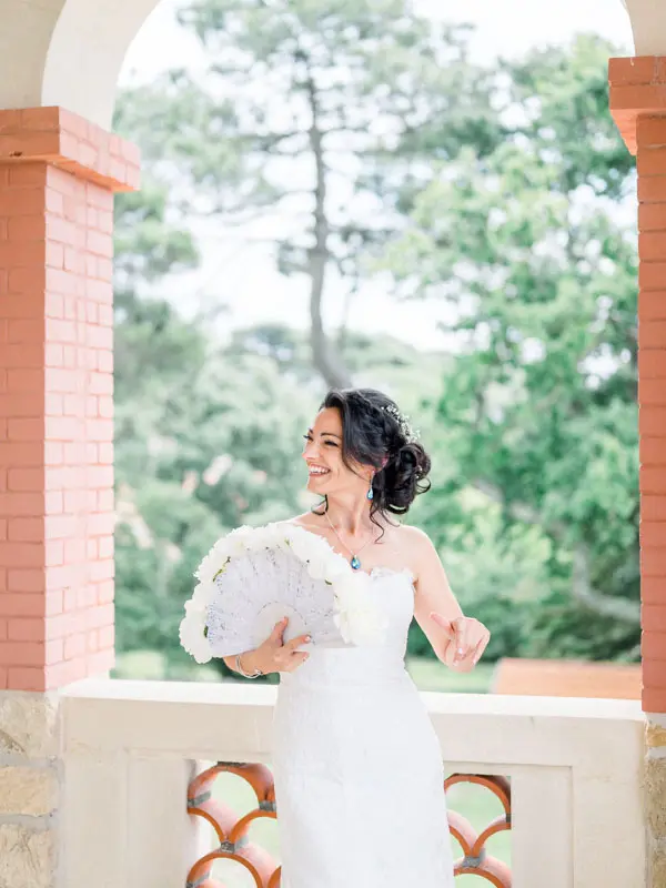 The smiling bride holding a white floral fan beneath the arcades
