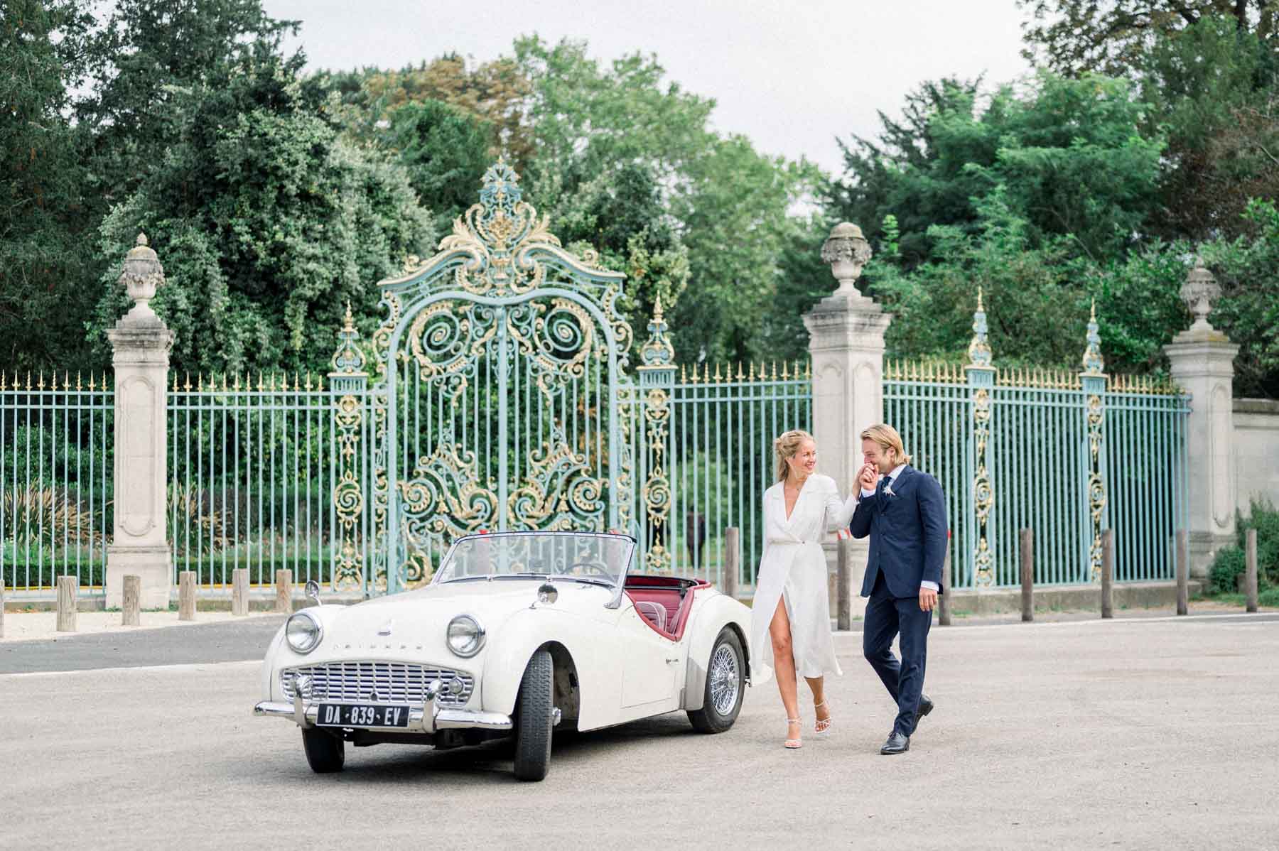 The couple walking past a gilded gate beside a white vintage convertible