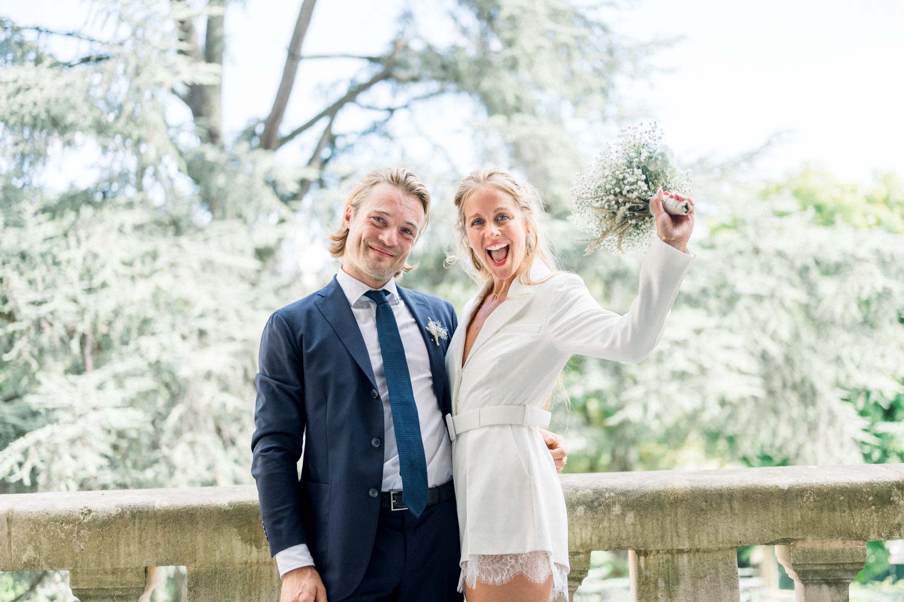 The bride raises her bouquet, laughing beside the groom on the town hall balcony