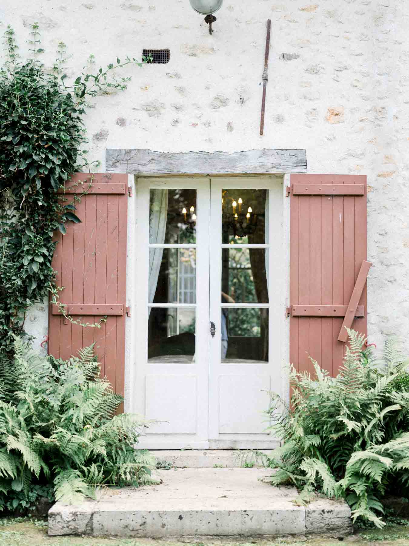 White French door framed by pink shutters and ferns at Moulin de Launoy