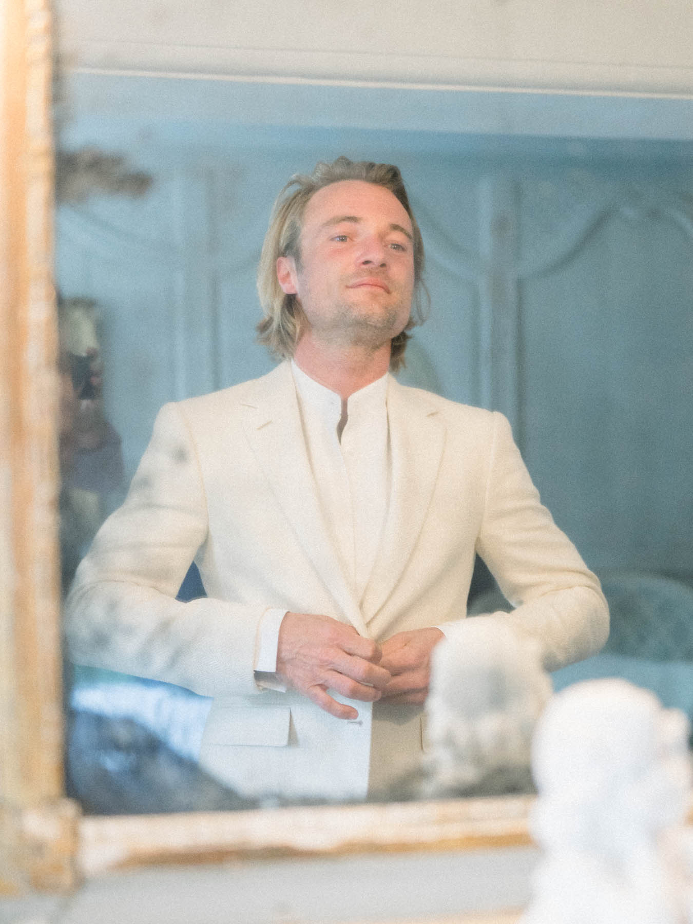 The groom in a cream jacket adjusting his outfit in a mirror while getting ready
