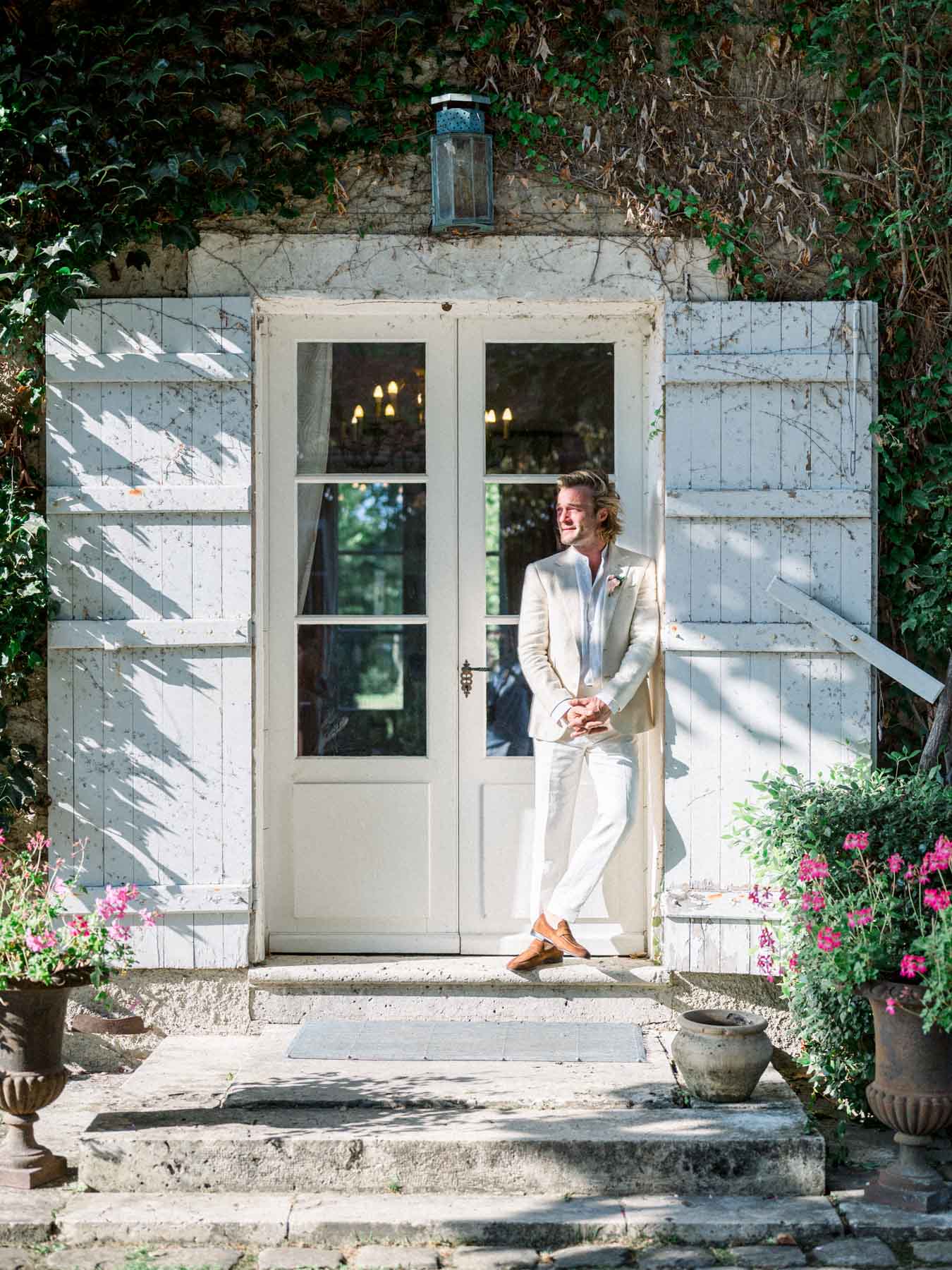 The groom in a cream suit waiting on the doorstep between white shutters