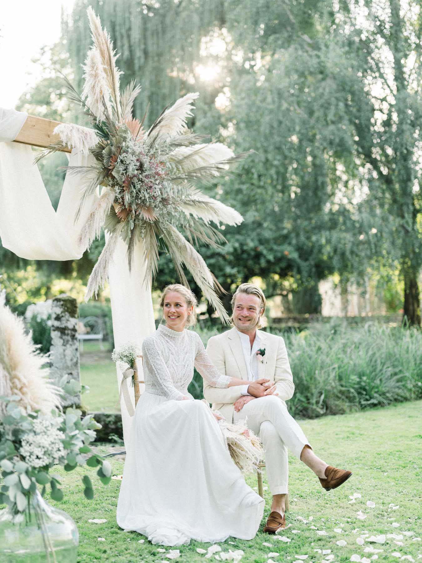 Virginie and Victor seated beneath the dried-flower arch during the outdoor ceremony