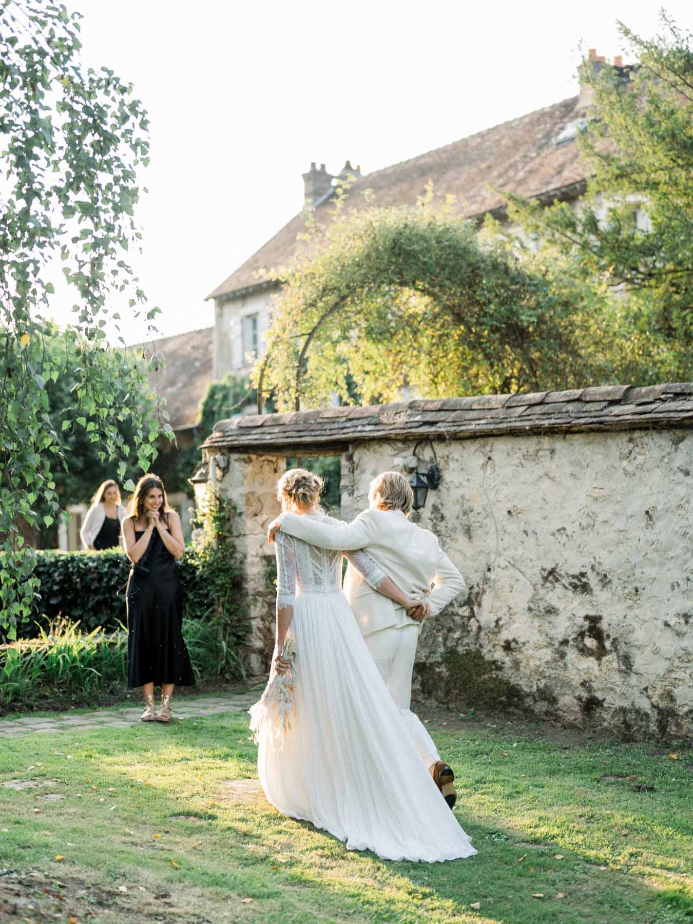 The couple embracing by a stone wall as a guest takes their picture